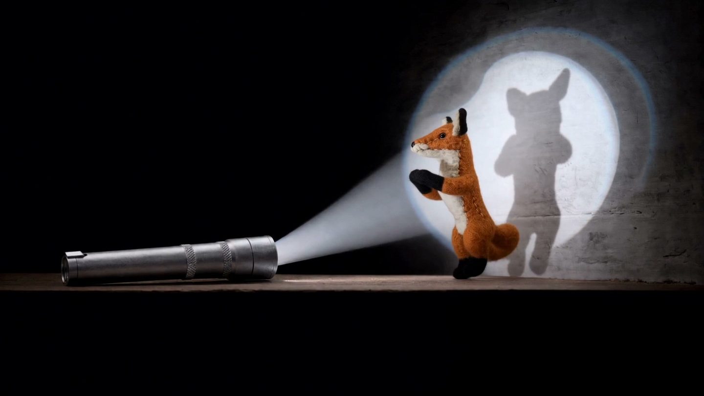 A felt fox stands in the beam of a pocket light, its shadow tall on the wall behind it.